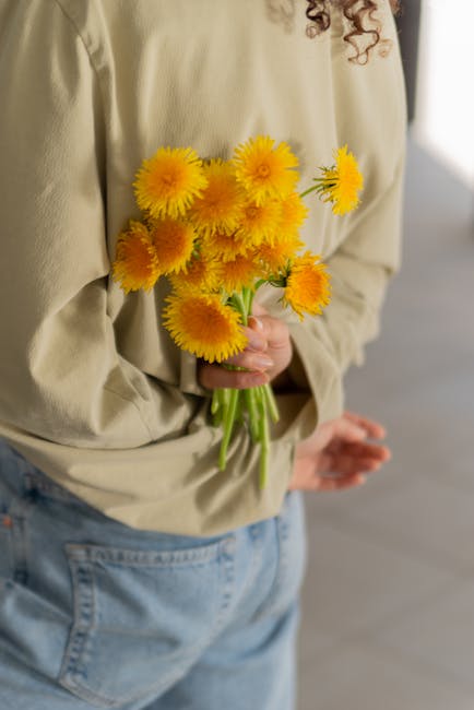 A person wearing a beige long-sleeve top and light blue jeans is holding a bouquet of bright yellow sunflower-like flowers with lush, textured petals and green stems. The flowers are arranged loosely,