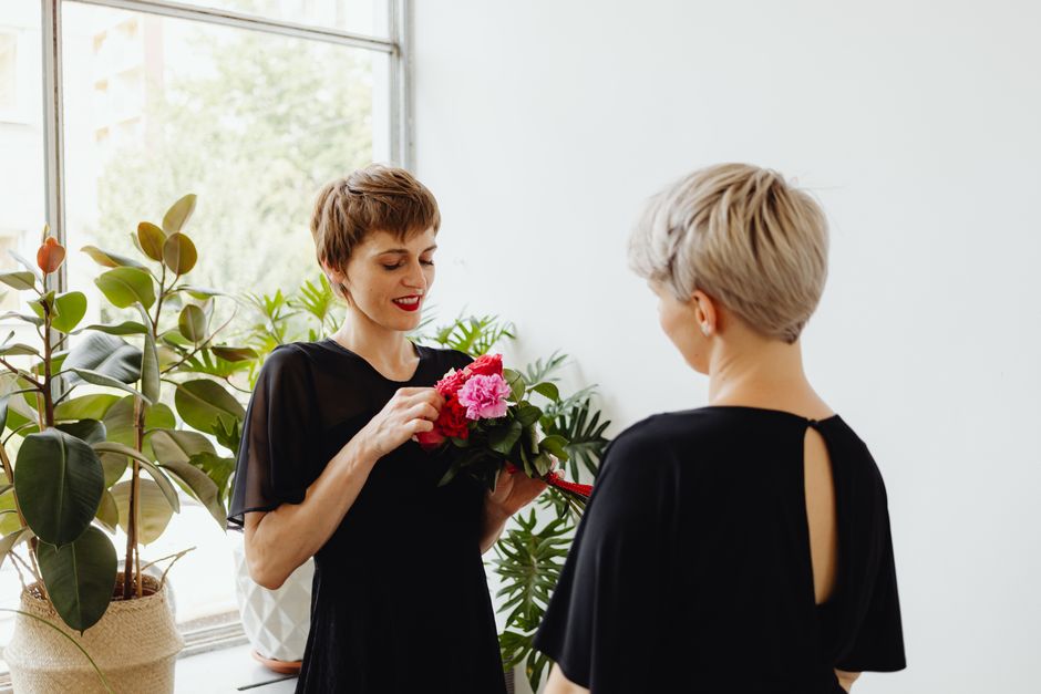 A woman with short brown hair and red lipstick holds a vibrant bouquet of pink peonies and dark green leaves, wrapped with a red ribbon, while smiling and engaging with another woman with short blond 
