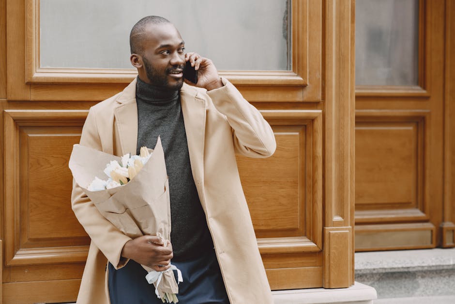 A smiling man with dark skin and a neatly trimmed beard is standing outdoors against a wooden-paneled wall, holding a bouquet of fresh white flowers wrapped in brown craft paper. He is dressed in a be