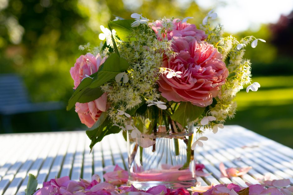 A floral bouquet arrangement featuring large, fully bloomed pink peonies with ruffled petals, complemented by delicate white blossoms and bright green leaves, all displayed in a clear glass vase. The 