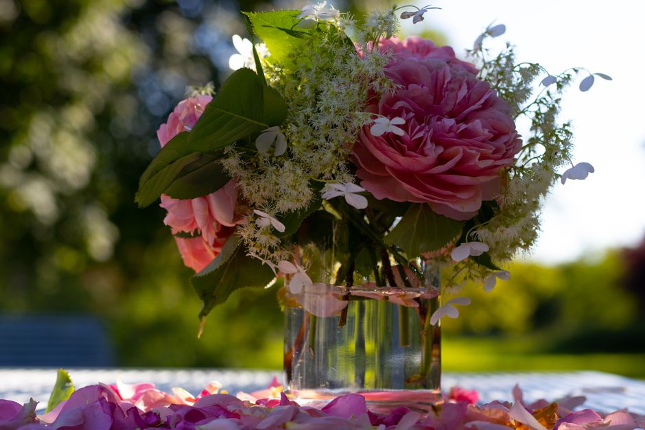 A close-up of a floral arrangement in a clear glass vase, featuring large pink peonies with soft, ruffled petals and fresh green leaves. The bouquet is complemented by smaller white blossoms and delic
